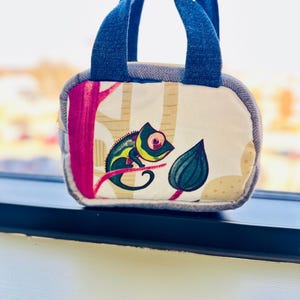 May include: A small, square tote bag with a denim blue handle and trim. The bag features a colorful print of a chameleon on a branch with a leaf. The background has a beige and pink tree design.