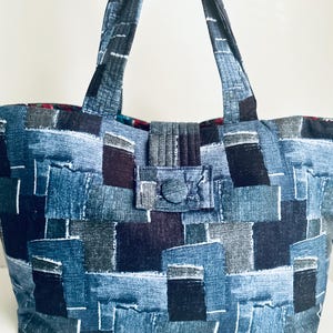 May include: A tote bag featuring a patchwork design of various blue, black, and grey rectangular fabric segments. The bag includes two fabric handles and a central tab closure secured with a matching fabric-covered button. The interior lining is visible.