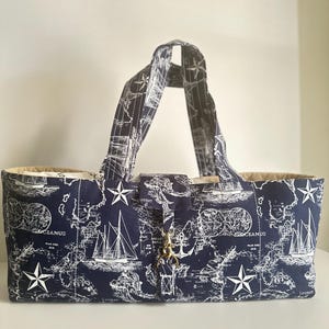 May include: A rectangular fabric tote bag featuring a dark navy blue background with a white nautical map print, including illustrations of sailboats, anchors, and compass stars. The bag has two fabric handles and a central tab with a gold metal clasp.