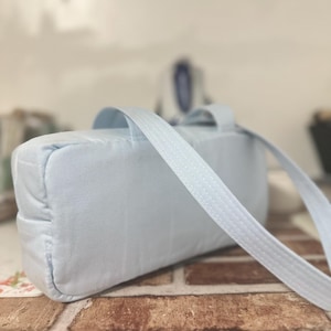 May include: A light blue rectangular bag with a matching strap. The bag is made of a soft, textured fabric and has a handle on top. The strap is attached to the sides of the bag and is designed to be worn over the shoulder.
