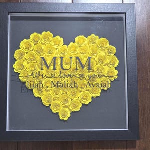 Könnte beinhalten: Gerahmtes Kunstwerk mit einem herzförmigen Arrangement aus gelben Papierrosen. Das Wort „MUM“ ist zentriert, darunter „We love you“ und Namen. Der Rahmen ist schwarz, der Hintergrund grau.