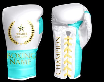 Puede incluir: Guantes de boxeo blancos y turquesa con detalles dorados. Un guante presenta una corona de laurel dorada con una estrella y las palabras "PREMIER SUPER FALCON". El otro guante tiene la palabra "CHAMPION" en oro.