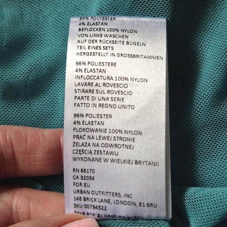 Puede incluir: Una etiqueta de ropa blanca con texto en varios idiomas, incluyendo ingl&eacute;s, alem&aacute;n, italiano y polaco. La etiqueta detalla la composici&oacute;n de la tela, las instrucciones de cuidado y la direcci&oacute;n de Urban Outfitters, Inc. en Londres.