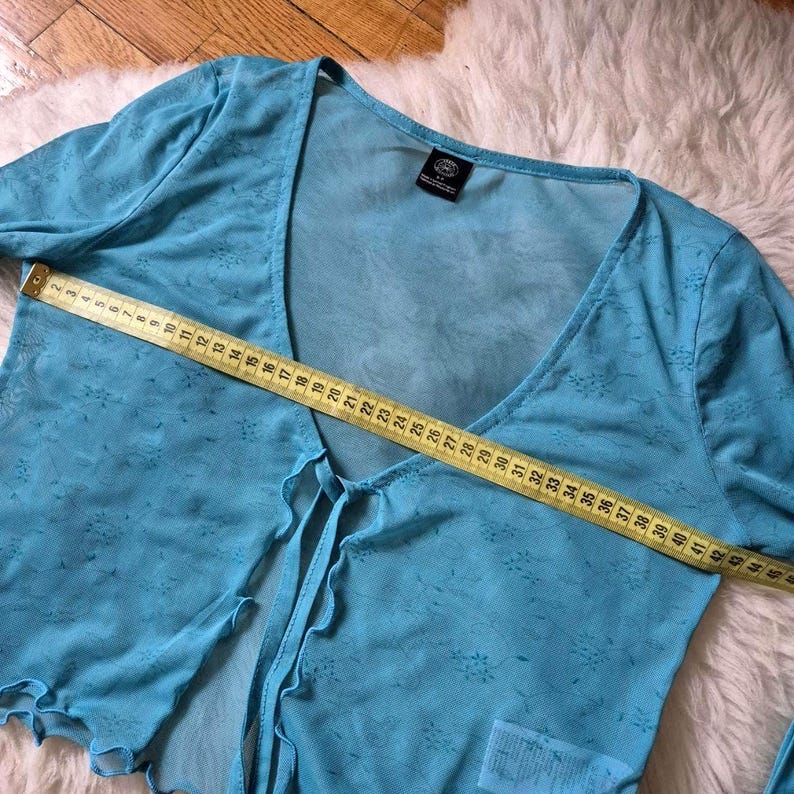 Puede incluir: Top de manga larga azul claro y transparente con cuello en V y cierre de lazo. El top tiene un estampado floral y se mide con una cinta m&eacute;trica amarilla. El top mide aproximadamente 109 cm de ancho.