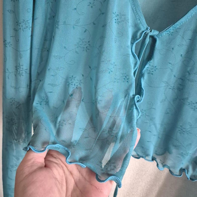 Puede incluir: Top transparente azul turquesa con mangas largas y un delicado estampado floral. El top presenta un cierre de lazo en el escote y bordes con volantes en las mangas y el dobladillo. La tela es ligera y semitransparente.