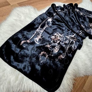 Puede incluir: Bufanda de terciopelo negro con un diseño floral en plata y oro rosa. La bufanda está doblada, mostrando el intrincado patrón y la suave textura. Se encuentra sobre una superficie blanca y esponjosa.