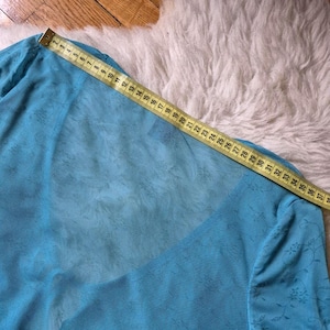 Puede incluir: Una blusa azul claro y transparente con mangas largas. Una cinta m&eacute;trica amarilla se coloca sobre la blusa, mostrando medidas en cent&iacute;metros. La blusa tiene un estampado floral en las mangas y est&aacute; hecha de una tela ligera.