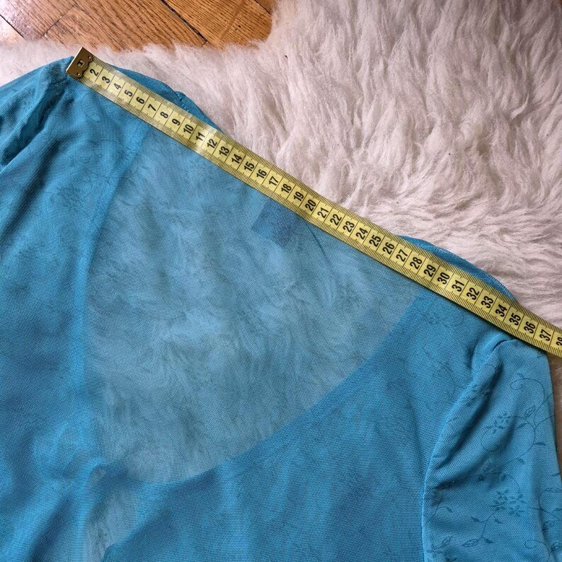 Puede incluir: Una blusa azul claro y transparente con mangas largas. Una cinta m&eacute;trica amarilla se coloca sobre la blusa, mostrando medidas en cent&iacute;metros. La blusa tiene un estampado floral en las mangas y est&aacute; hecha de una tela ligera.