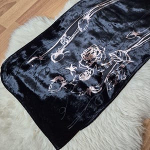 May include: A black velvet scarf with a floral design in shades of pink and silver. The scarf features a rose and leaf pattern, with a soft, textured appearance. The scarf is displayed on a white, fluffy surface.