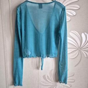Puede incluir: Top turquesa transparente de manga larga, con cuello en V y cord&oacute;n en la cintura. La tela tiene un sutil estampado floral. Los bordes de las mangas y el dobladillo son festoneados. Una prenda de moda para combinar o llevar sola.
