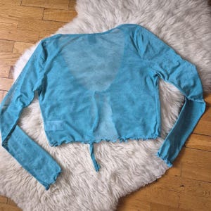 Puede incluir: Top corto transparente azul claro de manga larga con cierre de lazo. El top tiene un delicado estampado floral y un dobladillo con volantes. La prenda se muestra sobre una superficie blanca y esponjosa.