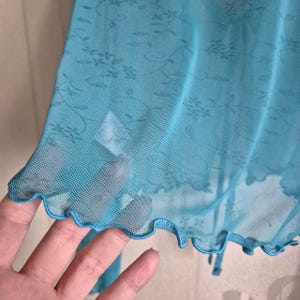 Puede incluir: Prenda transparente de color turquesa con un estampado floral. La tela tiene una textura delicada, similar a una red. El borde de la prenda es festoneado. El estilo general es delicado y femenino.