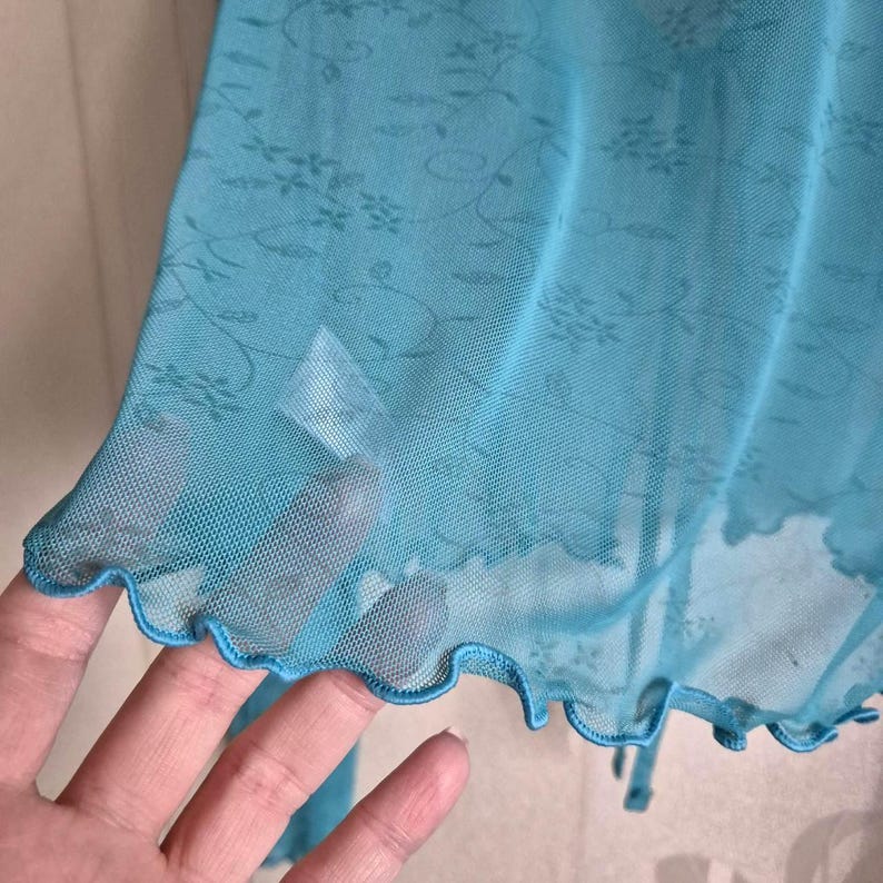 Puede incluir: Prenda transparente de color turquesa con un estampado floral. La tela tiene una textura delicada, similar a una red. El borde de la prenda es festoneado. El estilo general es delicado y femenino.