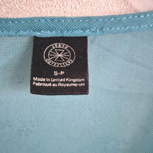 Puede incluir: Primer plano de una tela turquesa con una etiqueta negra. La etiqueta presenta el logotipo de Urban Outfitters y el texto "S-P Made in United Kingdom Fabriqu&eacute; au Royaume-Uni". La tela tiene una textura.