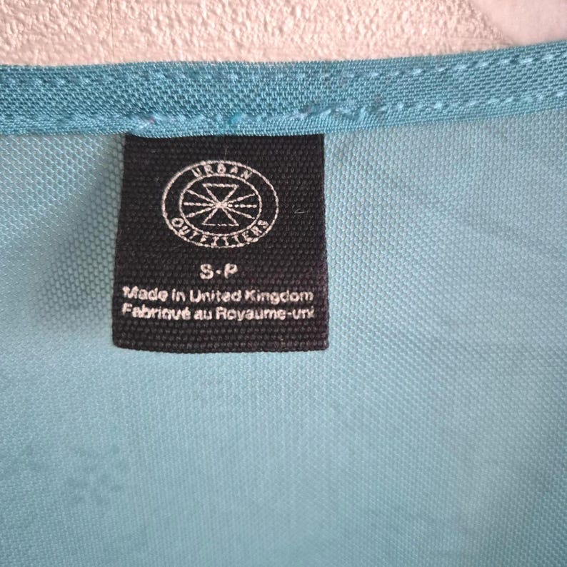 Puede incluir: Primer plano de una tela turquesa con una etiqueta negra. La etiqueta presenta el logotipo de Urban Outfitters y el texto "S-P Made in United Kingdom Fabriqu&eacute; au Royaume-Uni". La tela tiene una textura.