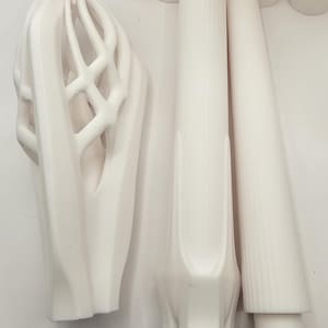 Op de afbeelding: Een verzameling witte 3D-geprinte onderdelen, waaronder een grote, holle, kooiachtige structuur met verticale ribben, een cilindrisch stuk met een uitlopende basis en een taps toelopende, geribbelde staaf, naast diverse kleine, eenvoudige cilindrische eindkappen.