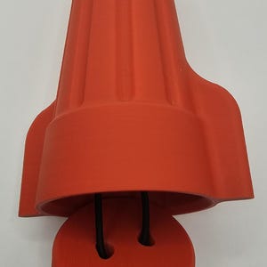 May include: Orange plastic wire connector with a black wire coming out of the top. The connector has a ribbed design and a rounded top. The connector is open and the bottom part is visible.