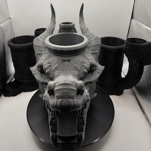 Puede incluir: Una escultura de cabeza de dragón gris con grandes cuernos y boca abierta, colocada sobre una base circular negra. Varias tazas negras con diseños intrincados están dispuestas alrededor de la escultura, creando un tema de fantasía.