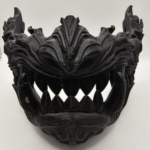 May include: A black mask with a detailed, fearsome design. The mask has sharp teeth, a wide open mouth, and elaborate, textured features. The overall aesthetic suggests a Japanese Oni or demon theme, suitable for cosplay or display.