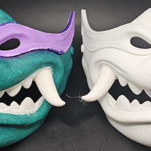 Puede incluir: Se muestran dos máscaras detalladas. Una es de color verde azulado con una cubierta para los ojos morada, dientes blancos y colmillos. La otra máscara es blanca con características similares. Ambas máscaras tienen una superficie texturizada y están diseñadas con ángulos agudos.