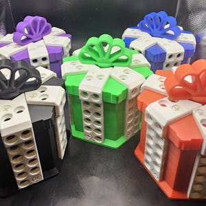 May include: Five hexagonal gift boxes in black, purple, green, blue, and orange, each featuring a white lid trim with circular indentations and a stylized bow on top. The boxes are constructed with a matte finish and have a geometric design.