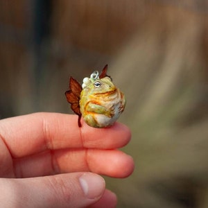 May include: A small, green and brown frog figurine with a white crown and brown wings. The figurine is sitting on a finger.