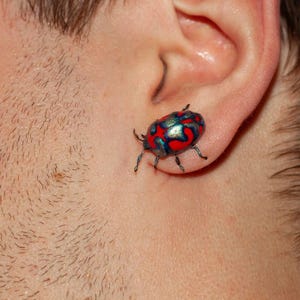 Op de afbeelding: Een oorsteker in de vorm van een kever met een metallic blauw en rood patroon op het schild. Het insectvormige sieraad heeft zes kleine pootjes aan de zijkanten, is bevestigd aan de oorlel en heeft een glanzende, reflecterende afwerking.