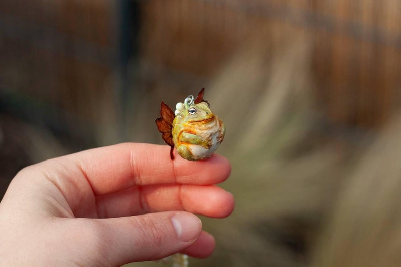 May include: A small, green and brown frog figurine with a white crown and brown wings. The figurine is sitting on a finger.