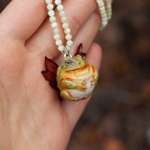May include: A necklace with a white pearl chain and a ceramic frog pendant. The frog is green, yellow, and brown with a white belly.