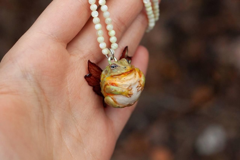 May include: A necklace with a white pearl chain and a ceramic frog pendant. The frog is green, yellow, and brown with a white belly.