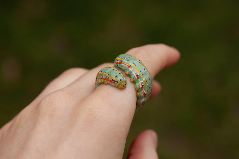 May include: A distinctive ring with a caterpillar design. The ring is made up of two parts, a smaller and a larger one, both in a light green colour with yellow, red, and white accents. The ring is worn on a finger.