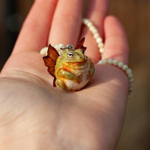 May include: A green and brown frog with wings, made of polymer clay, hangs from a white pearl necklace. The frog has a whimsical expression and is a handmade pendant.
