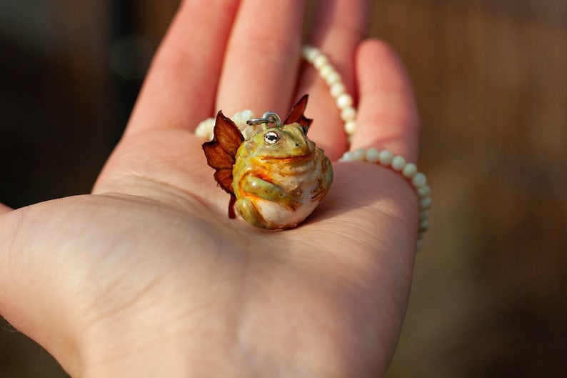 May include: A green and brown frog with wings, made of polymer clay, hangs from a white pearl necklace. The frog has a whimsical expression and is a handmade pendant.