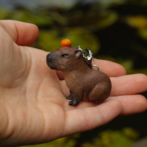 以下が含まれることがあります： 頭の上に小さなオレンジ色の果物を載せた、小さな茶色のカピバラのキーホルダー。キーホルダーは人の手に持たれています。