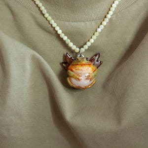 May include: A necklace with a white pearl chain and a ceramic frog pendant. The frog is painted in shades of green, brown, and orange.