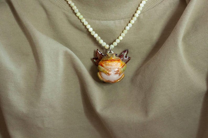 May include: A necklace with a white pearl chain and a ceramic frog pendant. The frog is painted in shades of green, brown, and orange.