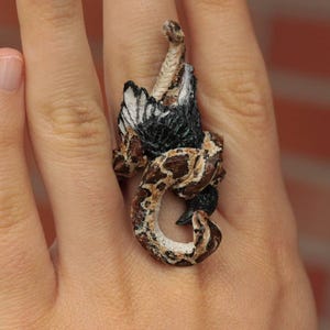 May include: A ring featuring a detailed sculpture of a snake wrapped around a bird. The snake is brown and black with a textured surface. The bird is black with white markings.
