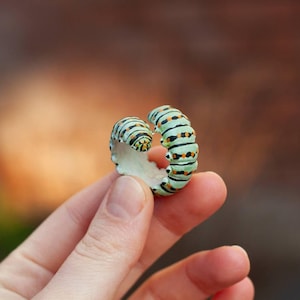 May include: A distinctive ring in the form of a caterpillar. The ring has a light green body with black and orange markings. The ring is held between two fingers against a blurred background.