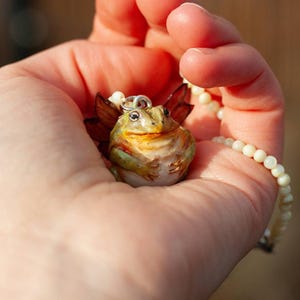 May include: A green and brown frog with wings, wearing a pearl necklace, is held in a hand. The frog has a wide smile and is made of ceramic.