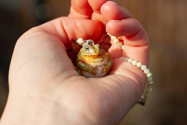 May include: A green and brown frog with wings, wearing a pearl necklace, is held in a hand. The frog has a wide smile and is made of ceramic.