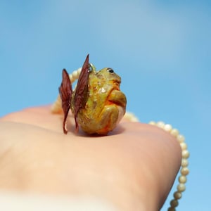 May include: A small, green and yellow frog figurine with brown wings, hanging from a white pearl necklace. The frog is wearing a pearl necklace and has a small, white pearl on its head.