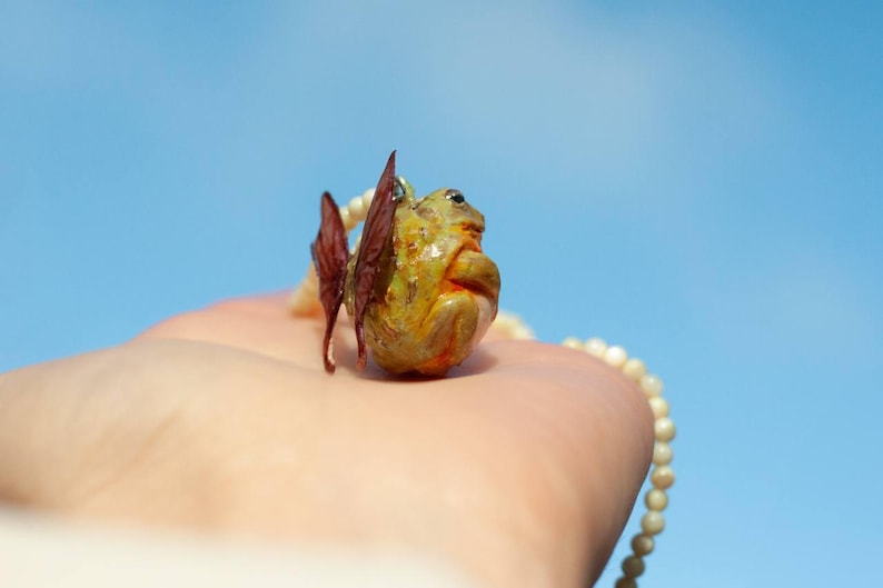 May include: A small, green and yellow frog figurine with brown wings, hanging from a white pearl necklace. The frog is wearing a pearl necklace and has a small, white pearl on its head.