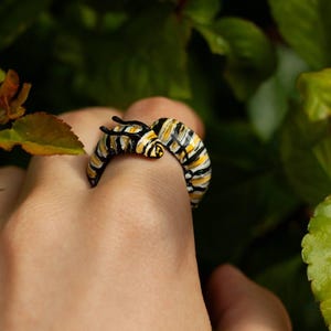 May include: A caterpillar-shaped ring with yellow, black, and white stripes. The ring's design includes two small antennae. The ring is set against a background of green leaves, offering a nature-inspired aesthetic.