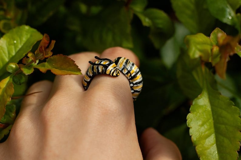 May include: A caterpillar-shaped ring with yellow, black, and white stripes. The ring's design includes two small antennae. The ring is set against a background of green leaves, offering a nature-inspired aesthetic.