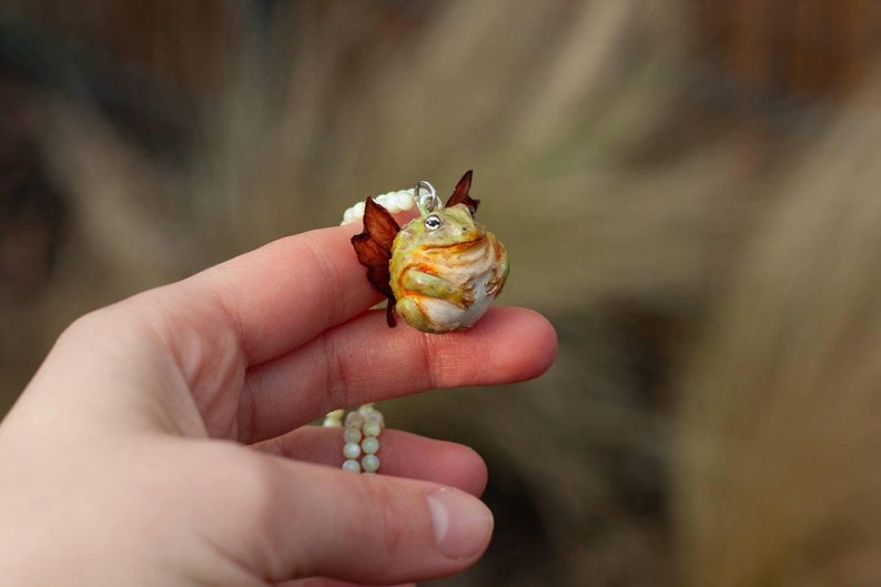 May include: A green and yellow frog with brown wings is attached to a white beaded necklace. The frog is sitting on a hand.