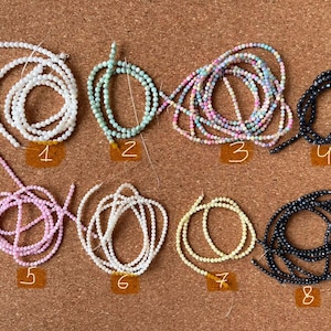 May include: Eight strands of beads, each in a different color and material, are laid out on a corkboard. The beads are arranged in two rows of four. The beads are white, light blue, pink, black, white, light yellow, and dark blue.