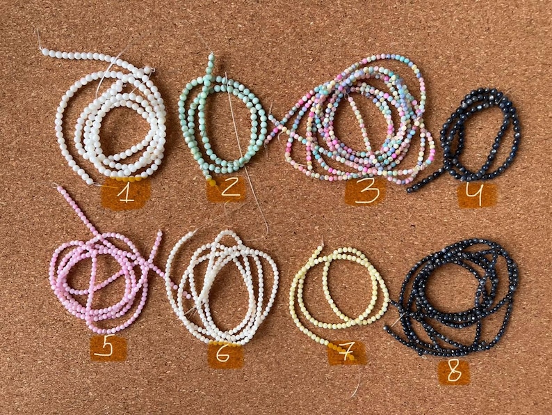 May include: Eight strands of beads, each in a different color and material, are laid out on a corkboard. The beads are arranged in two rows of four. The beads are white, light blue, pink, black, white, light yellow, and dark blue.