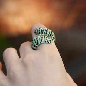 May include: A distinctive ring designed as two caterpillars. The ring has a light green hue with black stripes and small orange details. The ring is worn on a finger.