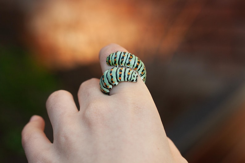 May include: A distinctive ring designed as two caterpillars. The ring has a light green hue with black stripes and small orange details. The ring is worn on a finger.