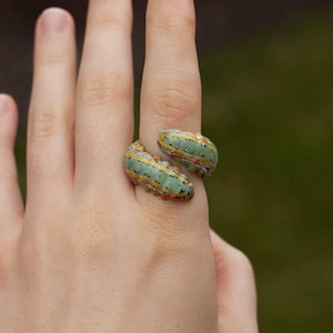 May include: A wrap-around ring shaped like a caterpillar. The ring features a light green body with horizontal yellow and orange stripes, accented by small black spots along the segments. The design mimics the appearance of a segmented larva.